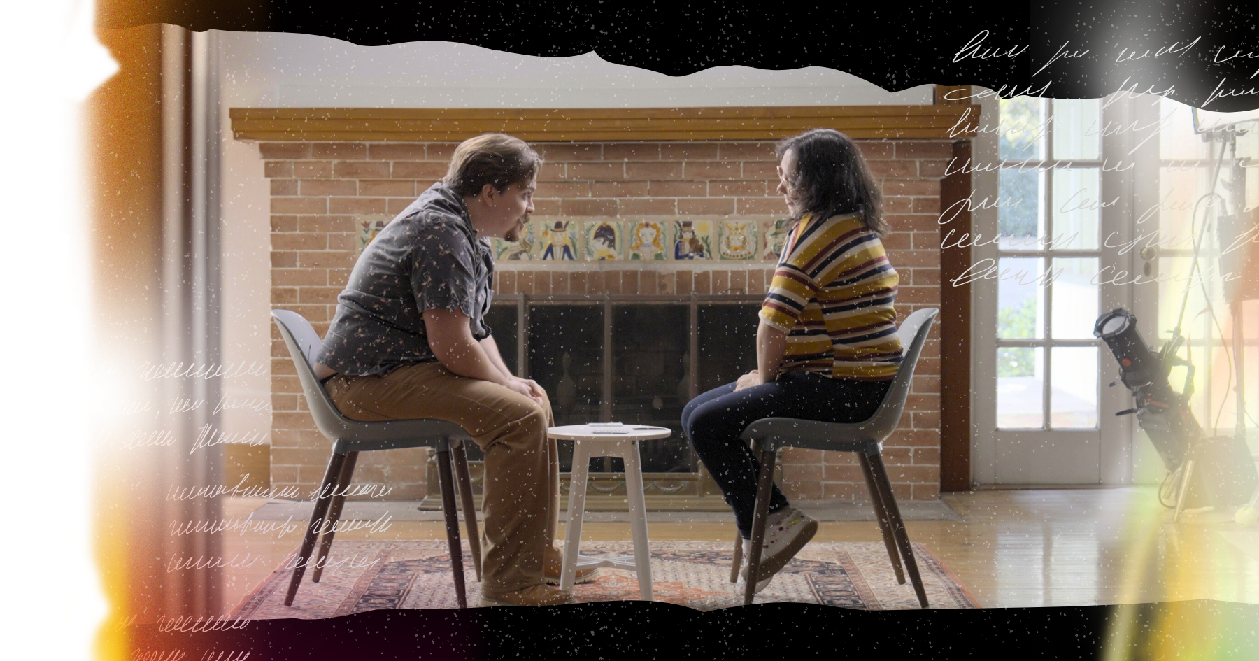 Image of a couple sitting face to face and smiling at each other, she wears glasses and he has a mustache, chimney behind them