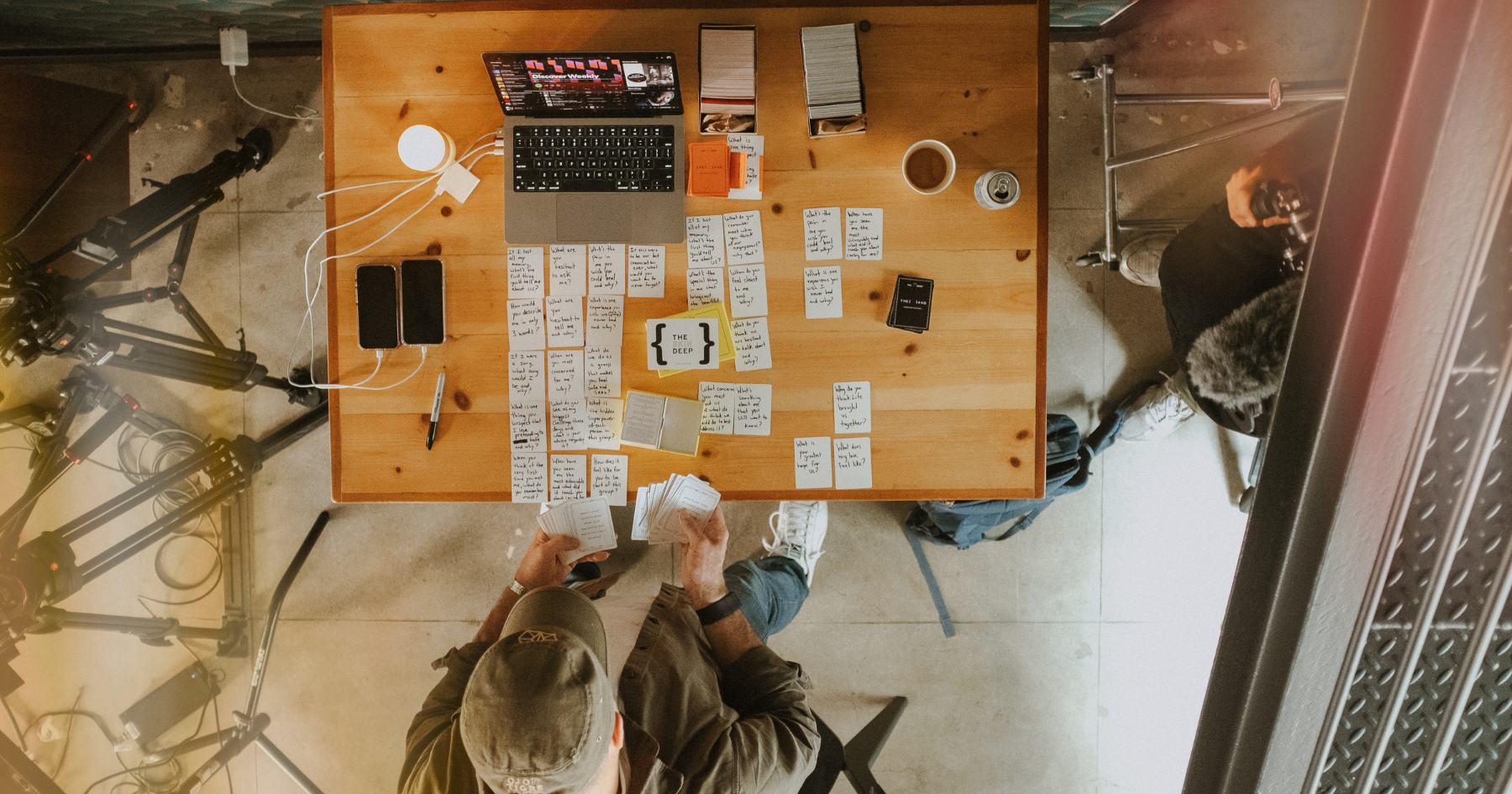 Picture of question cards on a wooden table, with a laptop, charging phones, a cup of coffee and a man