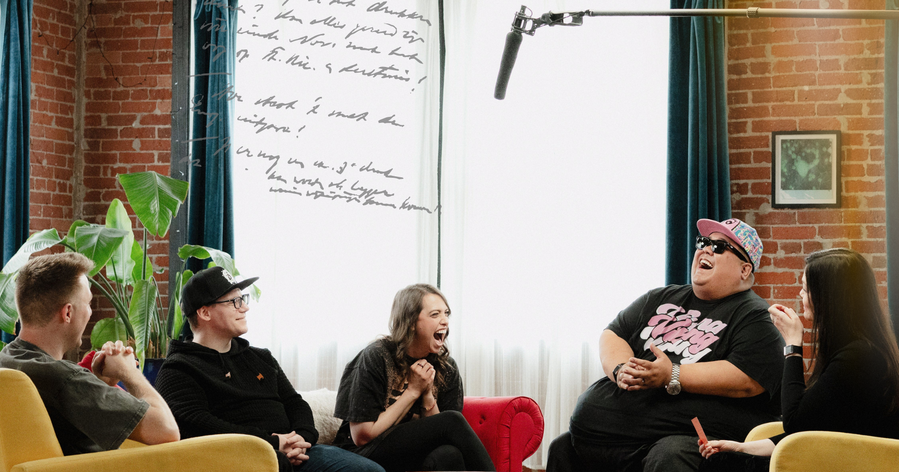 Picture of a group of 5 friends sitting down on yellow couches and laughing, a microphone above them