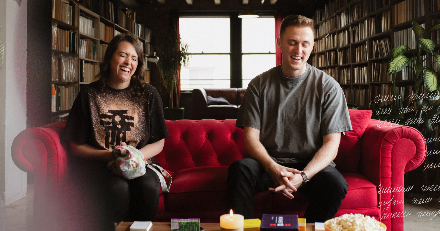 2 friends sitting down on a red couch and laughing together in front of a table with a candle, popcorn and card games, a library behind them