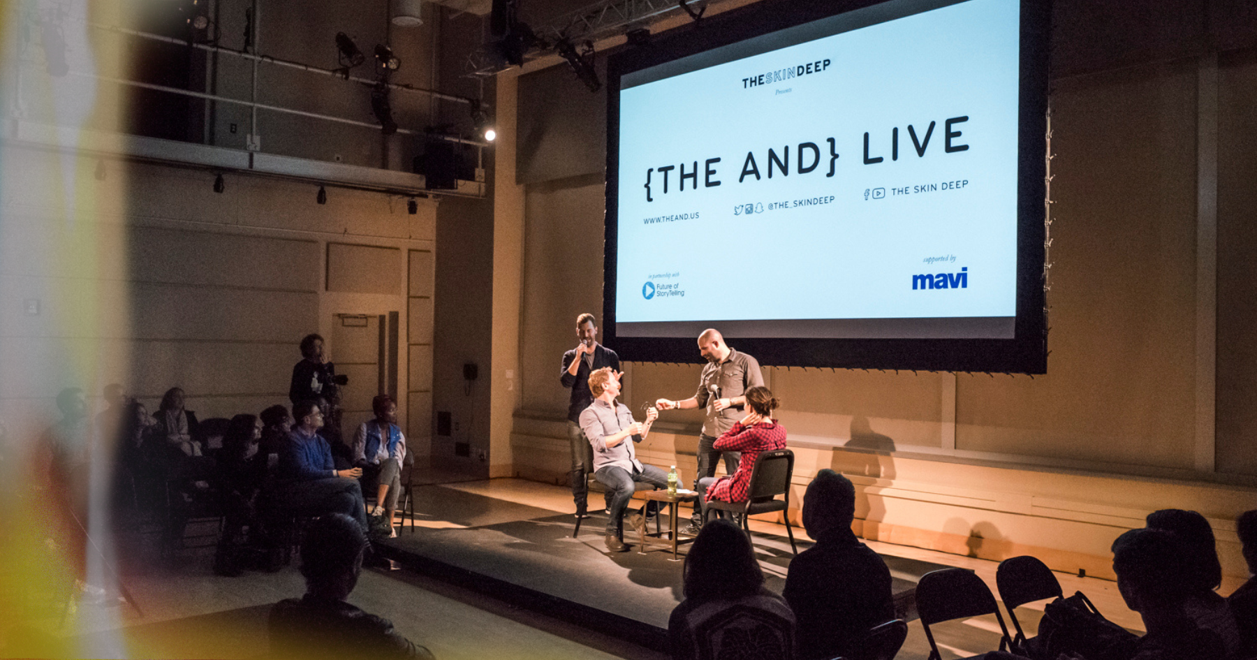 Picture of a stage full of people and two people seating face to face below a projector