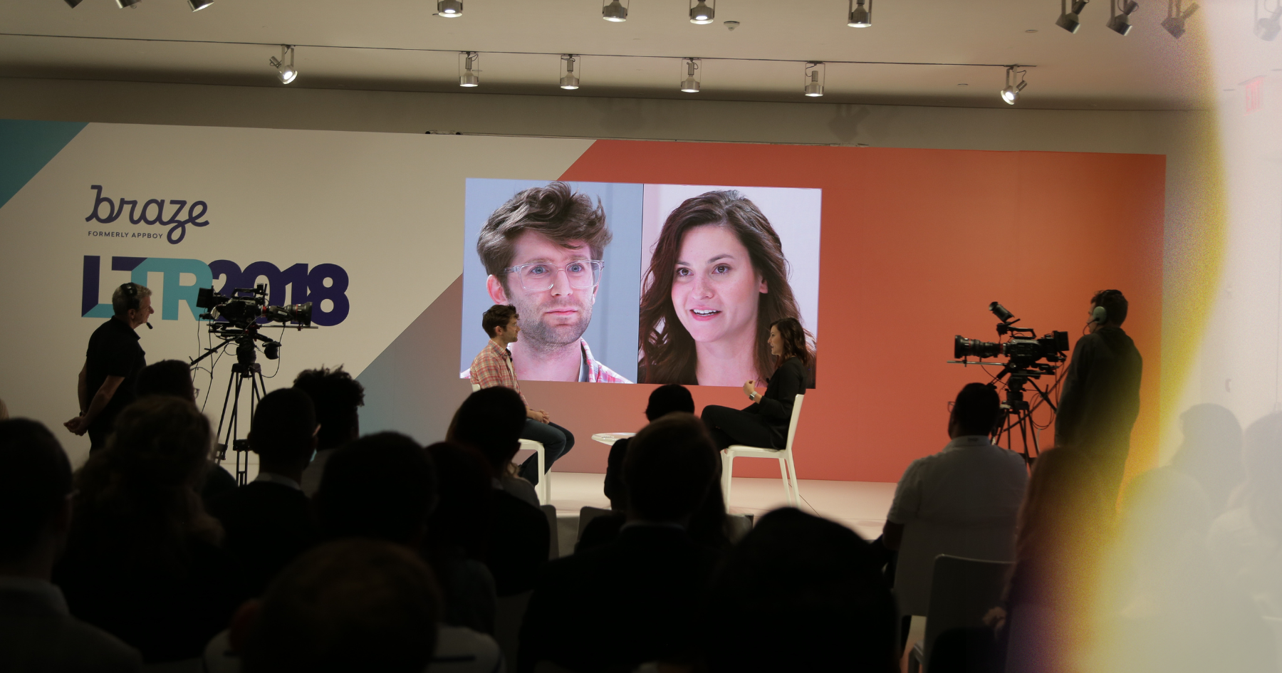 Picture of a room full of people and two people sitting face to face on a stage with their faces on the screen