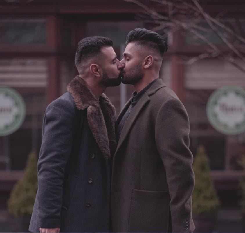 Picture of a South Asian Queer couple wearing coats and kissing on the street