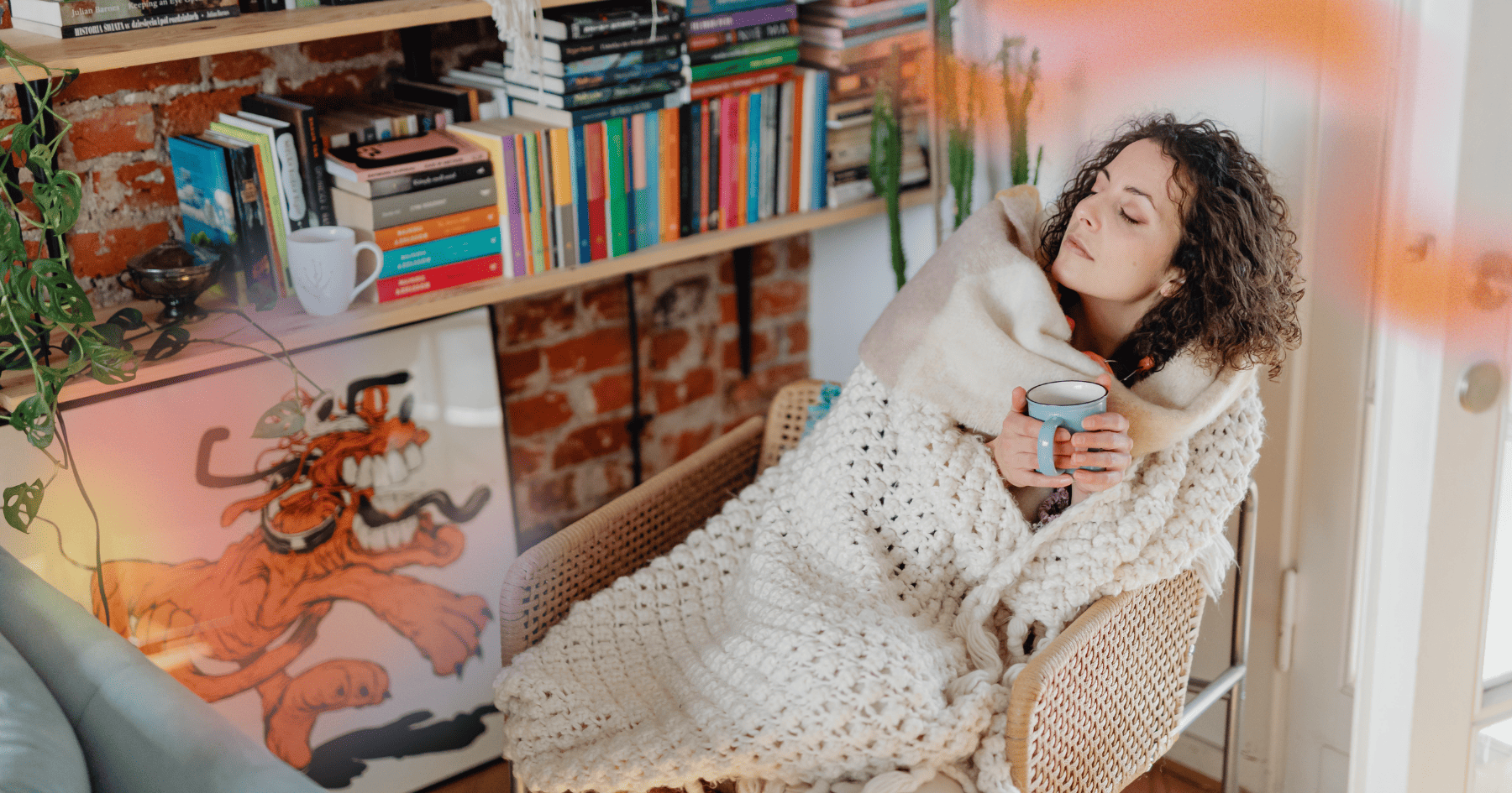 A woman sitting on a comfortable chair, drinking tea, wrapped inside a white blanket and next to a pile of books