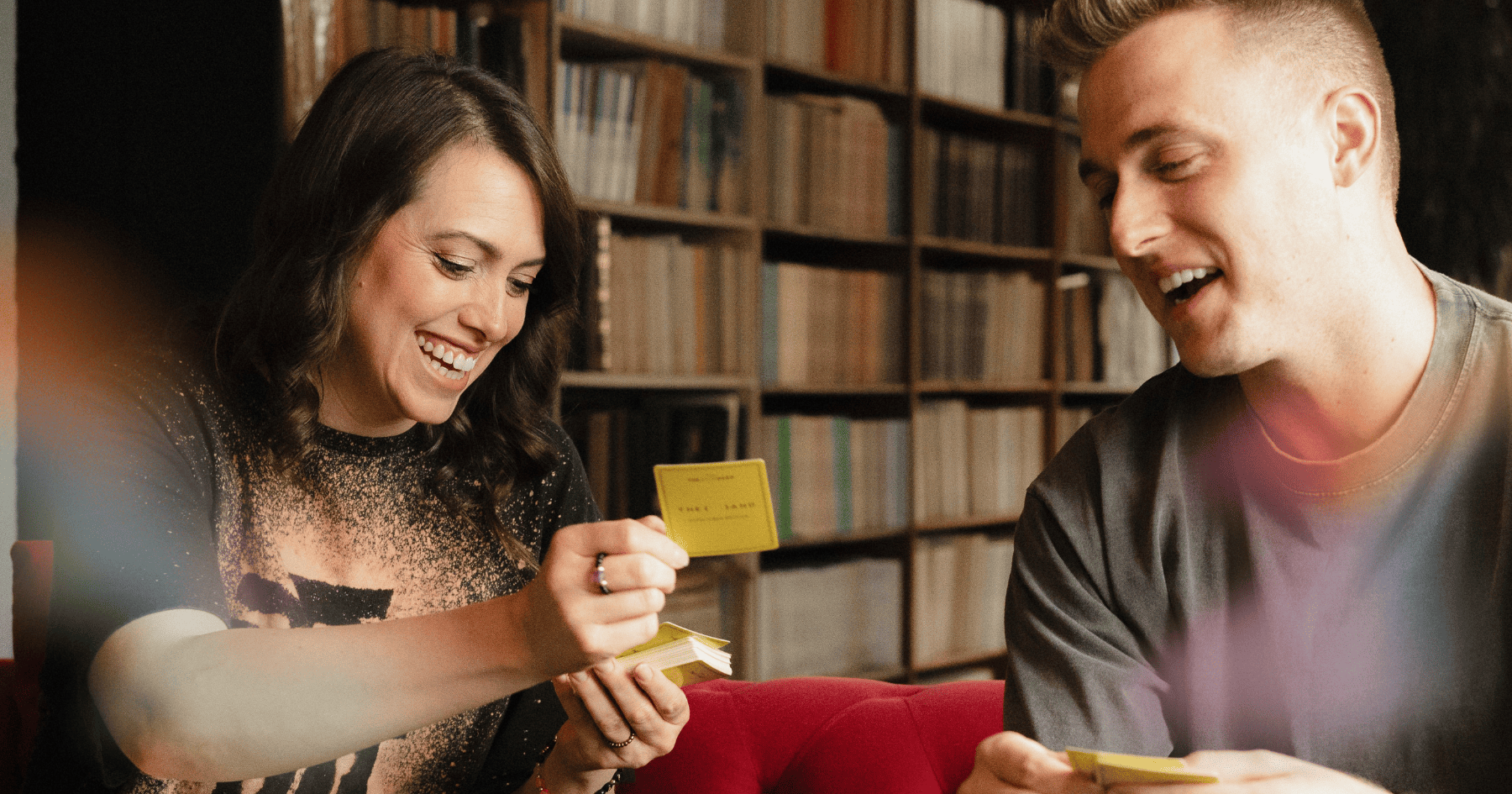 A man and woman from Sunny & The Black Pack playing {THE AND} Good Vibes card game sitting on a couch and laughing