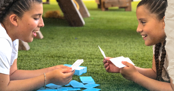 Picture of two young girls wearing braids and playing a card game laying on the grass and smiling
