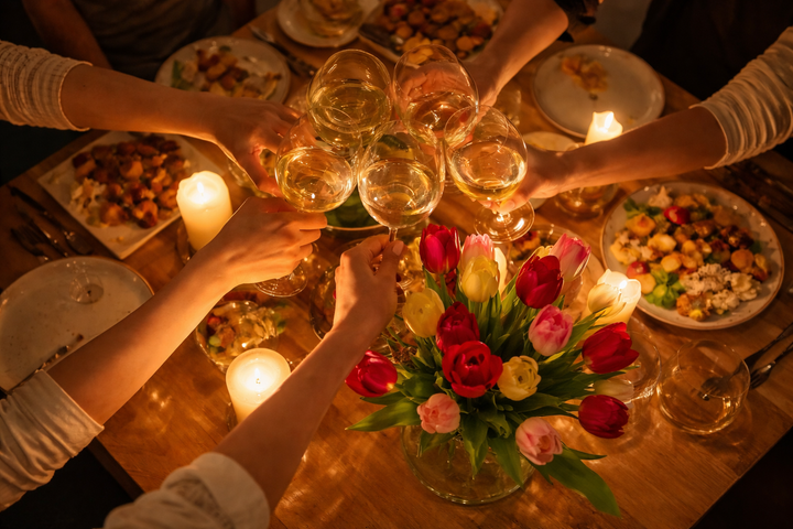 Overhead view of several people clinking wine glasses over a candlelit dinner table with tulips and shared plates of food.